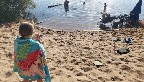 A child wrapped in a towel sits on a sandy beach, watching people swimming and relaxing by the water.