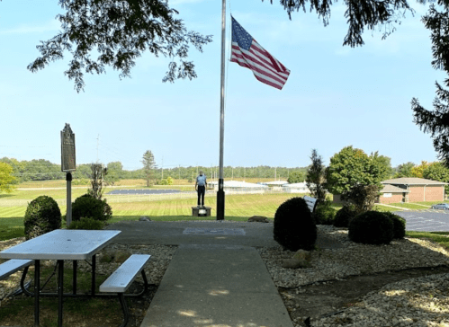 A person stands by a flagpole with an American flag, overlooking a grassy area and a building in the background.