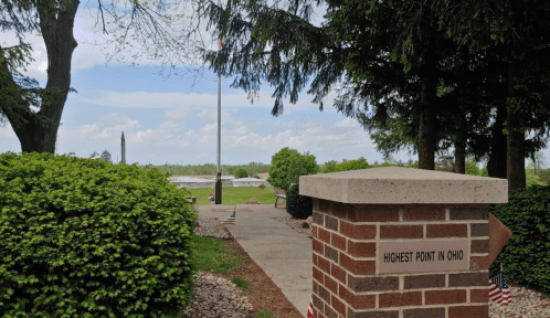 View of the Highest Point in Ohio marker, surrounded by greenery and flags, with a distant landscape under a blue sky.