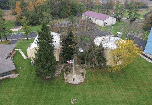Aerial view of a grassy area with two buildings, a flagpole, and trees, surrounded by autumn foliage.