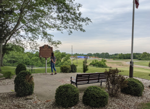 A person stands near a historical marker, overlooking a grassy area and a distant road, with trees and a cloudy sky.