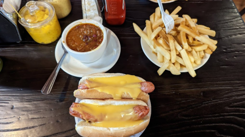 A plate with two cheese-covered hot dogs, a bowl of chili, and a side of golden French fries on a wooden table.