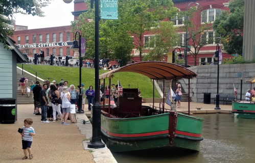 A green boat docked by a busy waterfront, with people gathered nearby and historic buildings in the background.