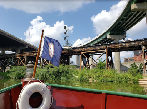 A boat with a Virginia flag on a river, surrounded by greenery and under a bridge with blue skies above.
