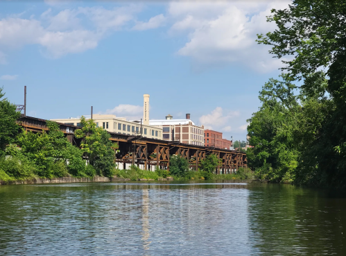 A calm river reflects a blue sky, with industrial buildings and lush greenery along the banks.