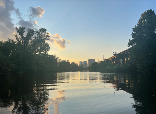 Sunset over a calm river, reflecting trees and city skyline, with clouds in a blue sky.
