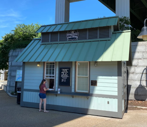 A small, light blue kiosk with a green roof under a bridge, featuring a menu board and a person at the window.