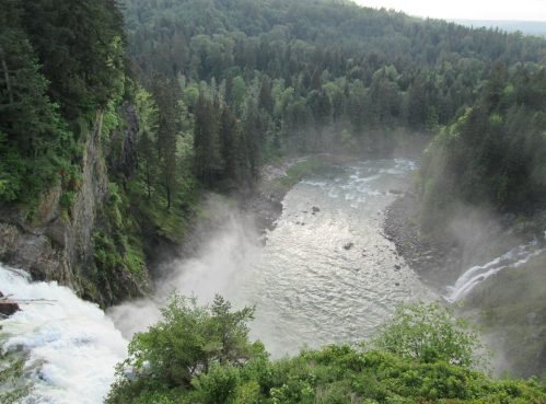Aerial view of a waterfall cascading into a river, surrounded by lush green trees and mist rising from the water.