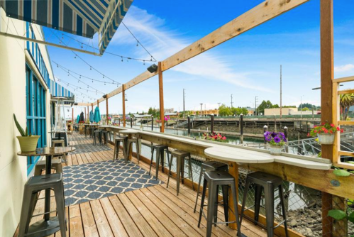 A waterfront patio with wooden decking, tables, and string lights, overlooking a calm waterway and blue sky.