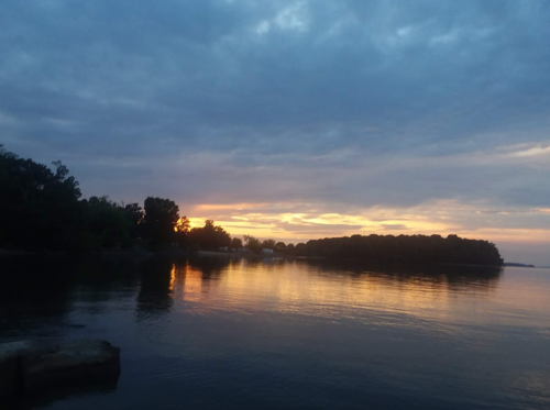 A serene lake at sunset, with colorful clouds reflecting on the water and trees lining the shore.