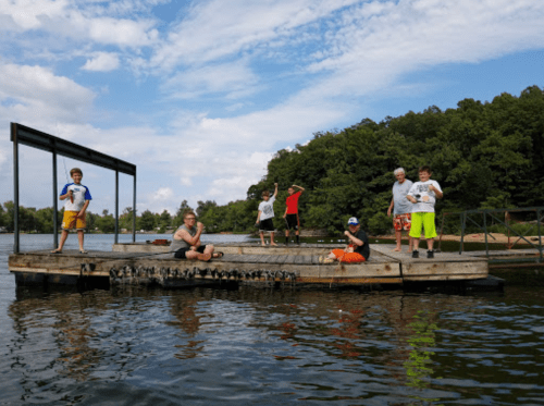A group of children and adults stand on a wooden dock by a lake, enjoying a sunny day outdoors.