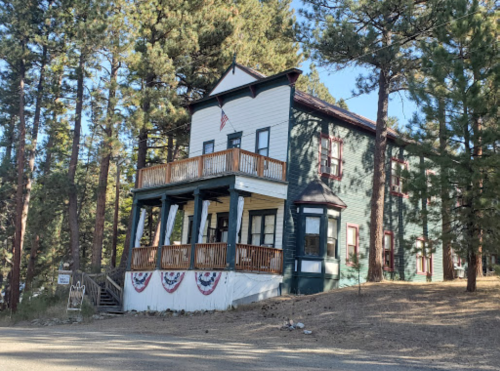 Historic green and white building with a porch, surrounded by tall pine trees and decorated with patriotic banners.