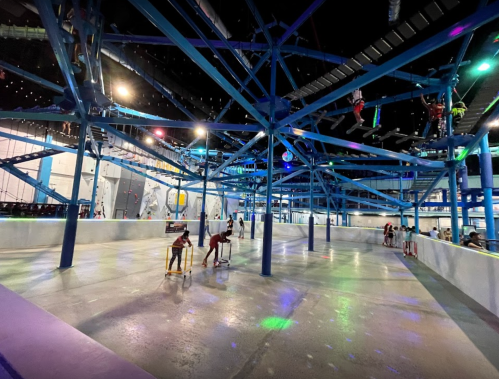 A spacious indoor activity area with blue metal structures, colorful lights, and people engaging in various activities.