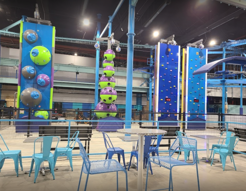 Indoor climbing gym featuring colorful climbing walls and seating area with blue chairs and tables.