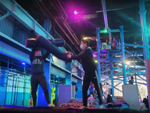 Two people in masks playfully spar with a large foam object in a colorful indoor gym with climbing structures.