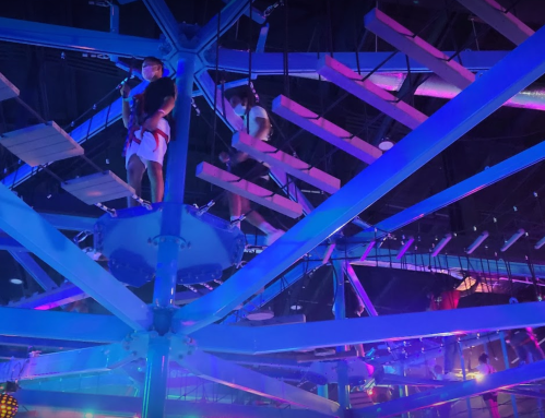 Two people navigate a colorful indoor ropes course, surrounded by vibrant blue and purple lighting.