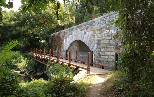 A wooden bridge leads to a stone archway surrounded by lush greenery and trees.