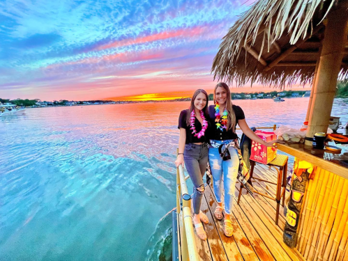 Two women in casual attire stand on a dock at sunset, wearing leis, with a vibrant sky and water in the background.