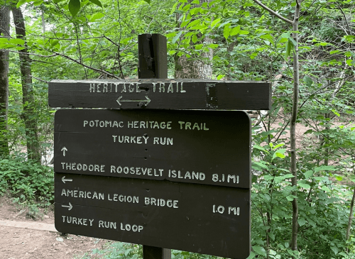 A wooden trail sign in a forest, indicating directions for the Potomac Heritage Trail and nearby locations.