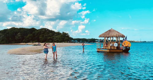 Two people wade in shallow water near a sandy shore, while a tiki boat floats nearby under a blue sky with clouds.
