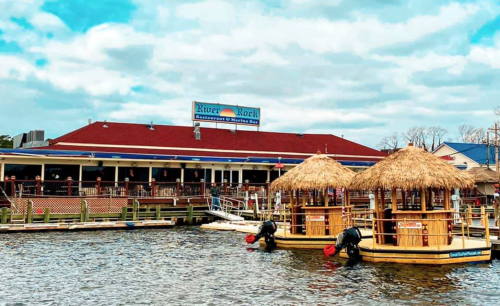 A waterfront restaurant with a red roof, featuring two tiki-style huts on the water and a cloudy sky above.