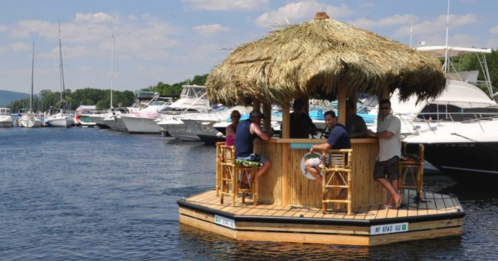 A tiki bar on a floating platform with people enjoying drinks, surrounded by boats on a calm waterway.