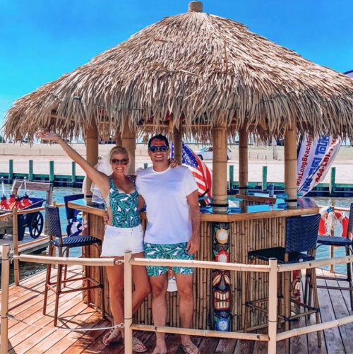 A couple poses in front of a tiki bar on a sunny day, wearing matching tropical outfits.