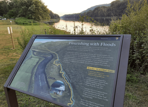 A sign titled "Flourishing with Floods" near a calm river, surrounded by greenery and trees in the background.