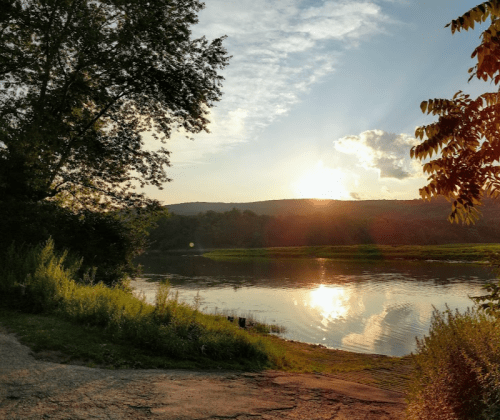 A serene river scene at sunset, with trees and hills reflecting in the water under a partly cloudy sky.