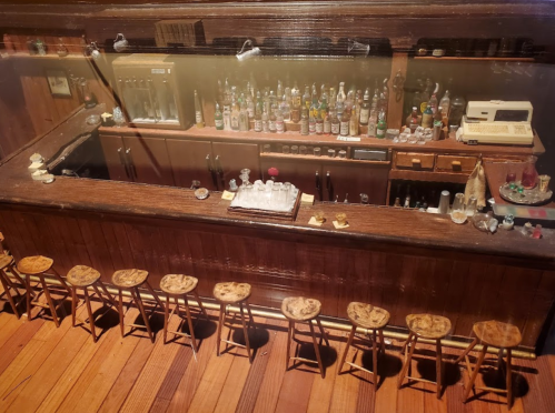 A detailed miniature bar scene with a wooden counter, stools, and shelves filled with various bottles.