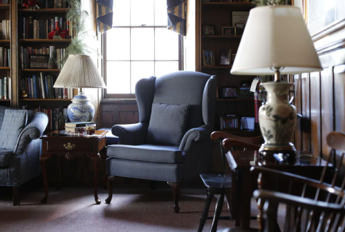 Cozy library scene featuring a blue armchair, wooden furniture, and bookshelves filled with books. Soft lighting from lamps.