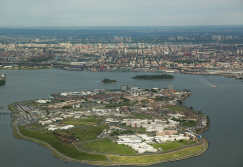 Aerial view of an island with buildings, surrounded by water and a city skyline in the background.