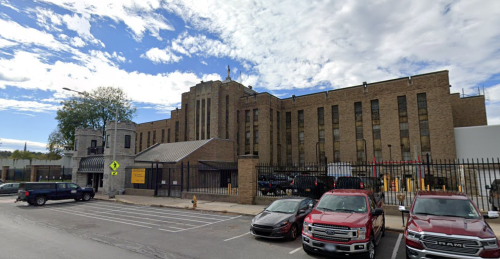 A large brick building with a distinctive architectural style, surrounded by a parking lot and vehicles.