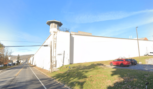 A large, white prison building with a guard tower, viewed from a road with parked cars and trees nearby.