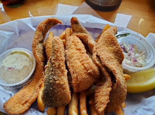 A plate of crispy fried fish fillets served with French fries, two dipping sauces, and a lemon wedge.