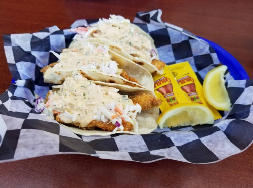 Three fish tacos topped with creamy sauce and coleslaw, served with lemon wedges and two packets of hot sauce.