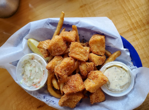 A basket of golden fried fish bites with fries, served with coleslaw and two dipping sauces on the side.