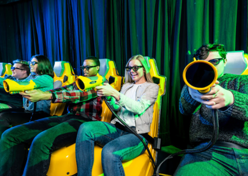 Five people in 3D glasses sit in yellow seats, aiming toy guns at a screen in a colorful, immersive gaming environment.