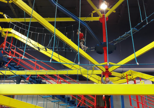 Two people navigate a colorful indoor ropes course with bright yellow and red structures.