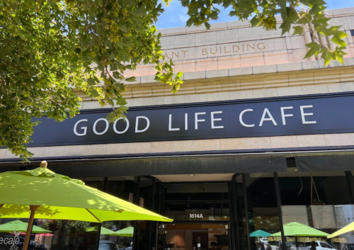 Sign for "Good Life Cafe" on a building, with green umbrellas in front and trees partially framing the view.