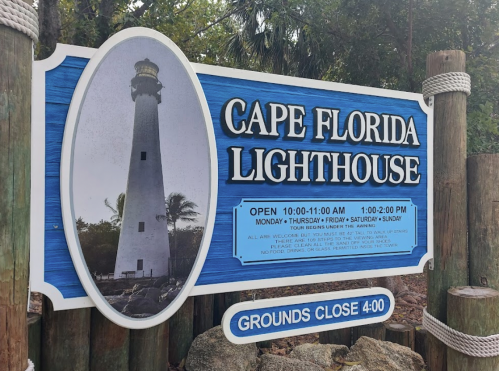 Sign for Cape Florida Lighthouse with hours of operation and a photo of the lighthouse. Grounds close at 4:00 PM.