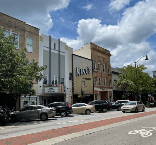 Street view of a downtown area featuring shops, trees, and parked cars under a partly cloudy sky.