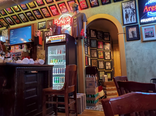 A cozy bar interior with framed posters, a drink fridge, and wooden chairs, creating a vibrant, eclectic atmosphere.