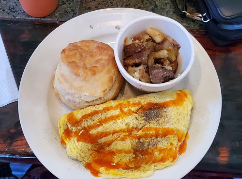 A plate with an omelette drizzled with sauce, a biscuit, and a side of seasoned potatoes.