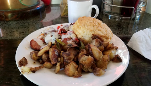A plate of breakfast featuring crispy potatoes, sausage, a biscuit, and a side of fruit with yogurt. Coffee in the background.
