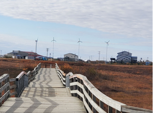 A wooden walkway leads through a grassy area towards buildings and wind turbines under a cloudy sky.