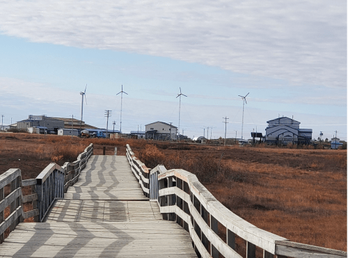 A wooden walkway leads through a grassy area towards buildings and wind turbines under a cloudy sky.