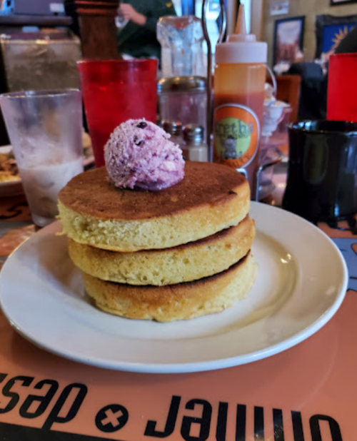 Three fluffy pancakes stacked on a plate, topped with a scoop of purple berry ice cream, surrounded by drinks and condiments.