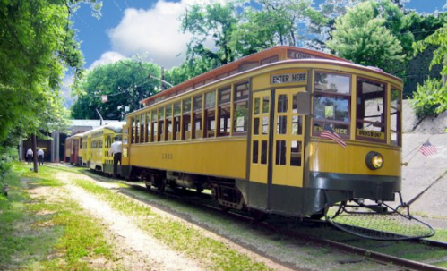 A vintage yellow streetcar parked on a green, tree-lined track under a blue sky.