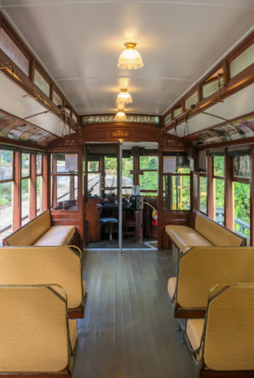 Interior of a vintage tram, featuring wooden benches, warm lighting, and large windows with a view of greenery outside.